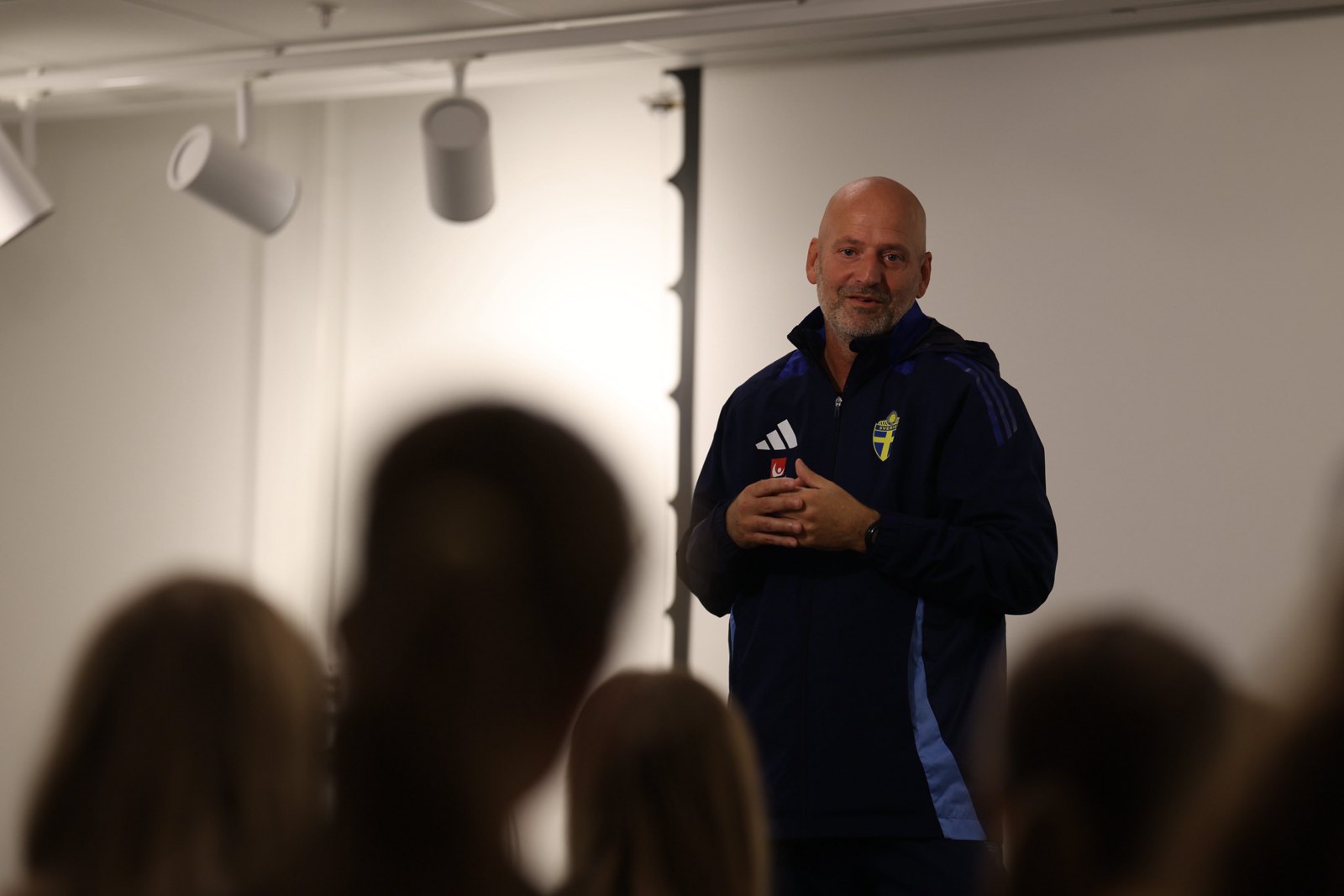 A coach in a Swedish national-team jacket speaking, with audience members in the foreground.